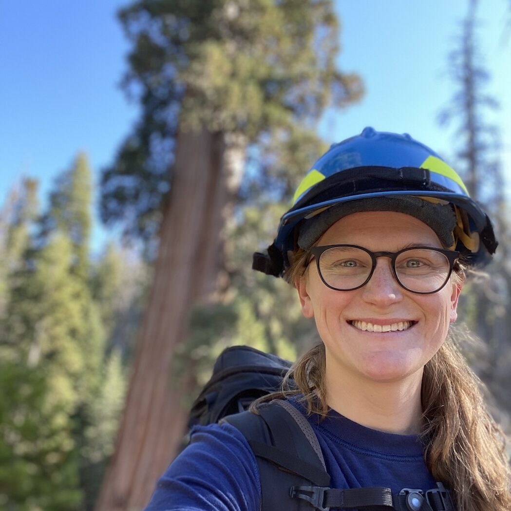 A woman wearing a hard hat and eyeglasses smiles in front of a giant sequoia tree.