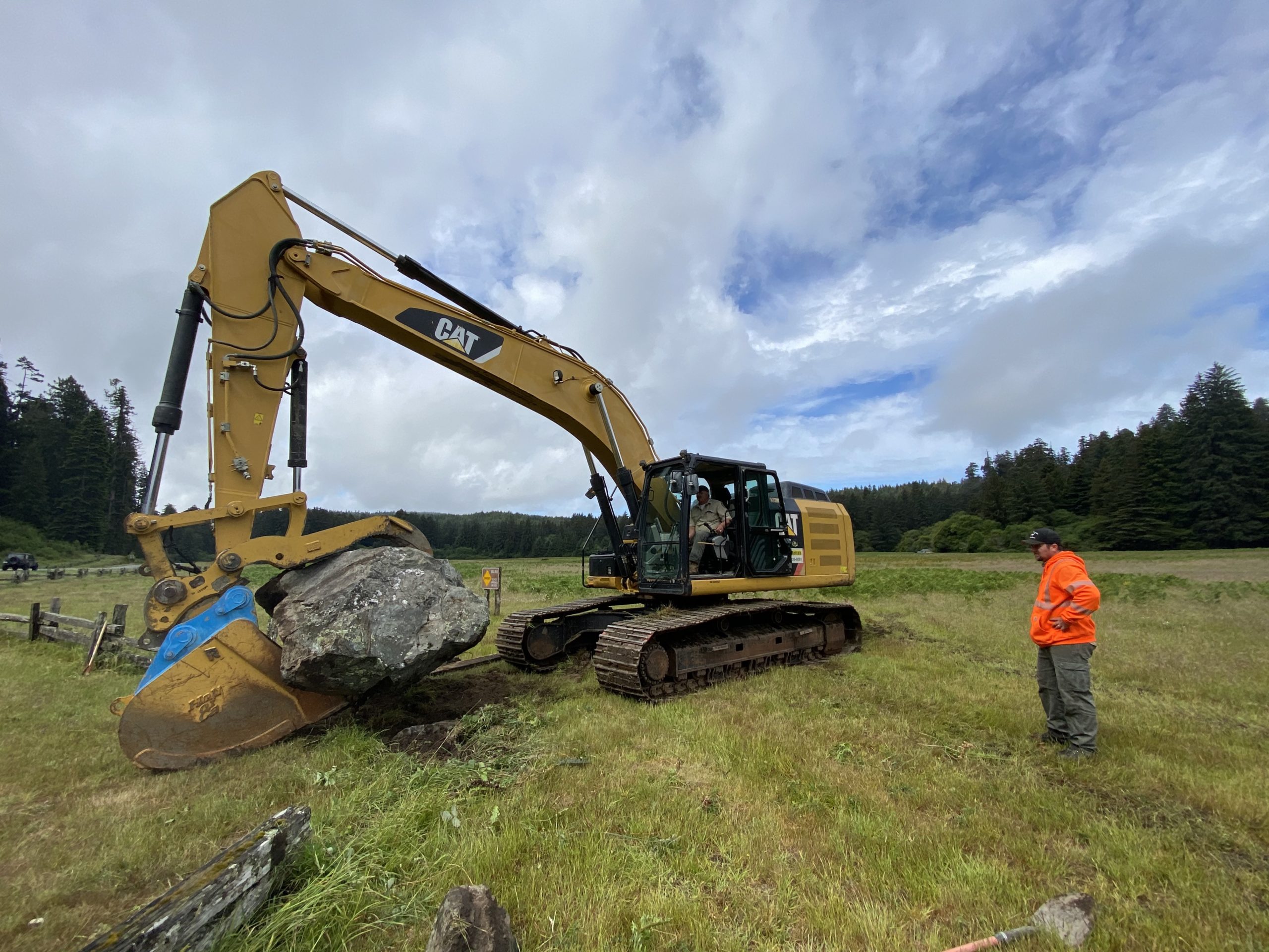 a large crane holds a massive rock as a man in an orange hi-viz jacket watches