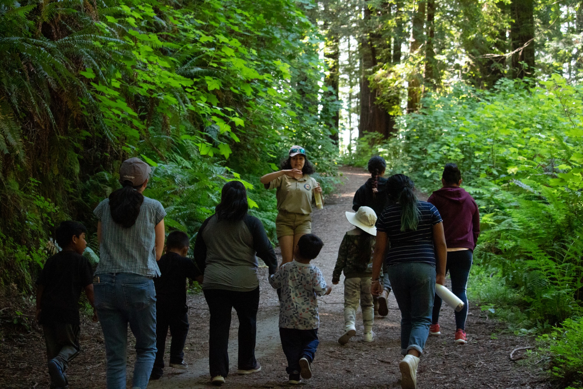 several people follow a park ranger who is walking backwards and speaking through a trail in a forested area