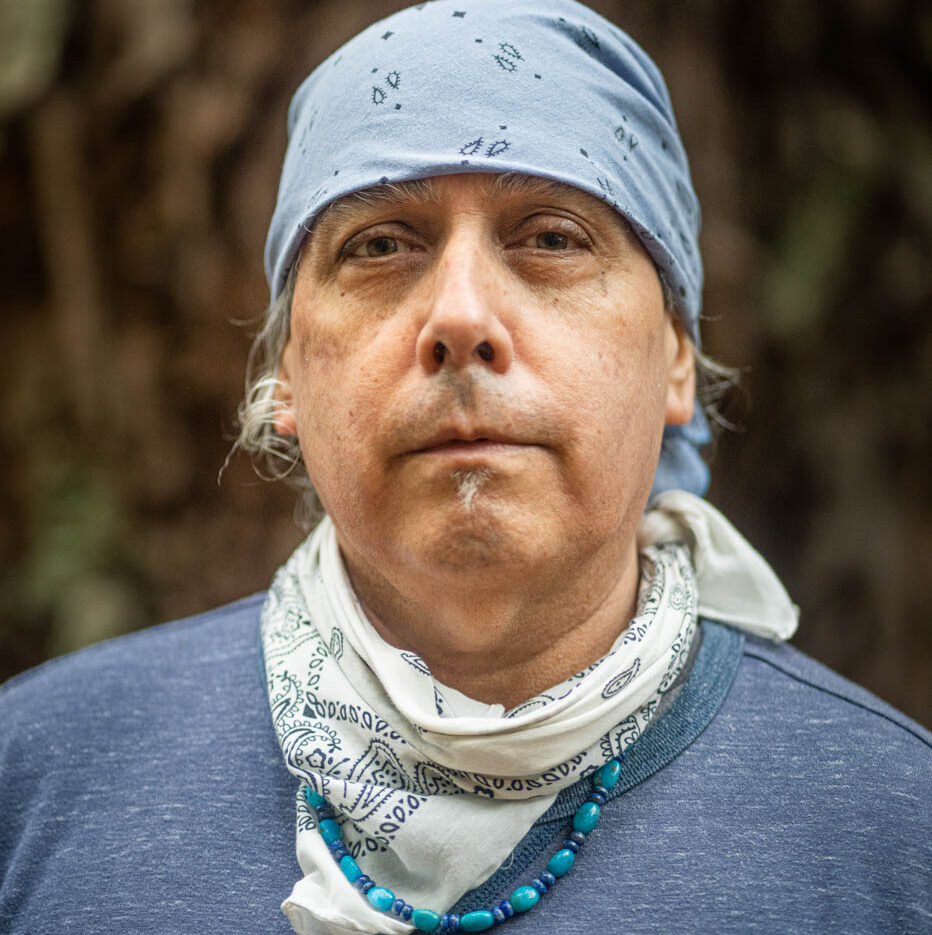 An indigenous man of mid-age wearing a blue bandana head-covering, white bandana neckerchief, a beaded necklace, and a crew-neck shirt.
