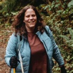 a person walks with a walking stick and a dog leashed to them through a forested trail