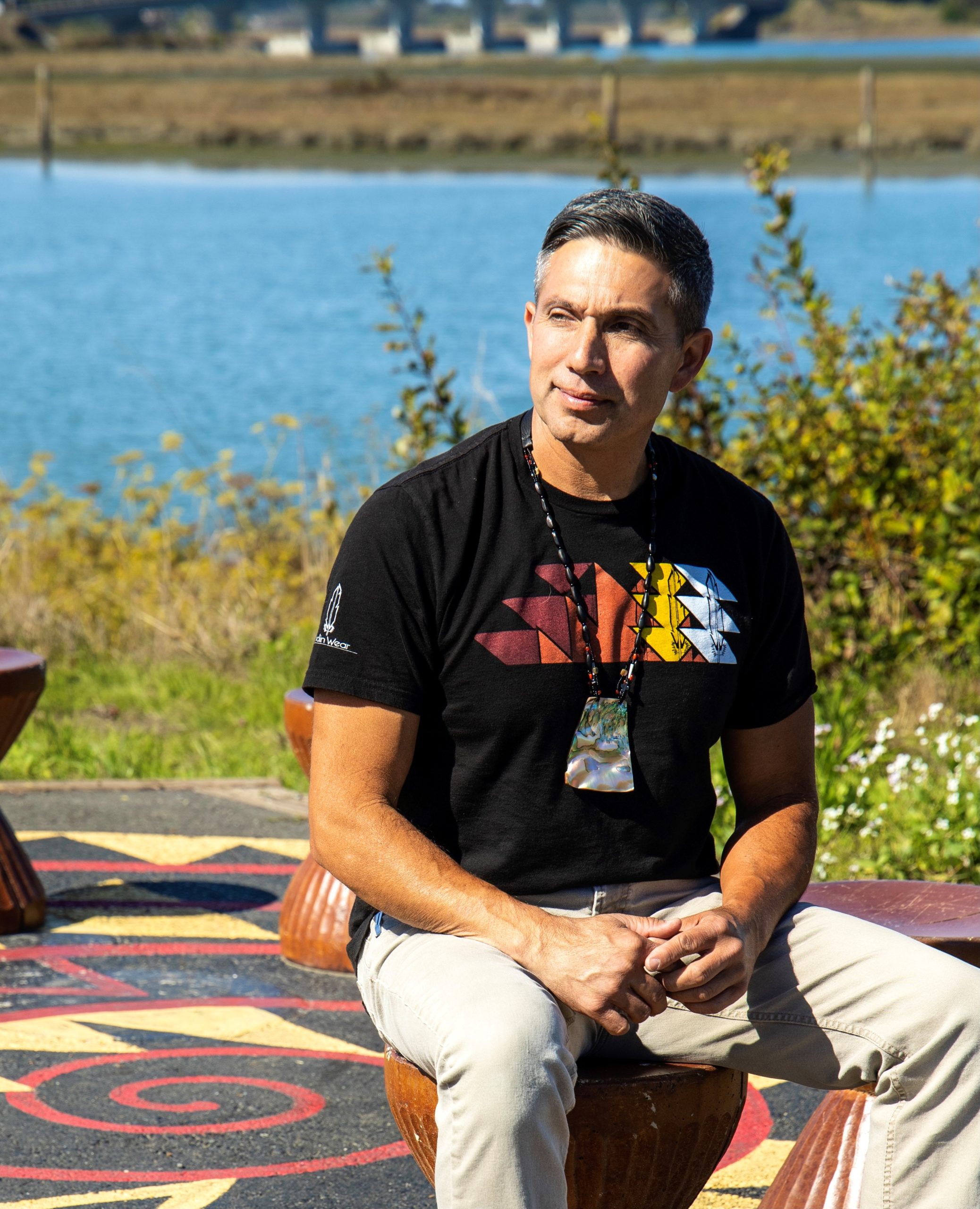 a man stares off toward the left while sitting on a small stool. behind him, there is a water body and some plants