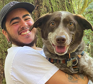 Jai Ledesma, a nonbinary transmasculine person of color with piercings and facial hair smiling and holding a very happy mixed breed dog