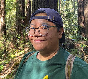 Portrait of Sam Mercado outdoors, a nonbinary AAPI person of color wearing a backwards blue hat, round glasses, and a green shirt.