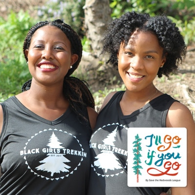 Two black women smiling, one with long braided hair and the other with chin length curls, wearing black tank tops with a circular logo featuring a tree and the words "Black Girls Trekkin"