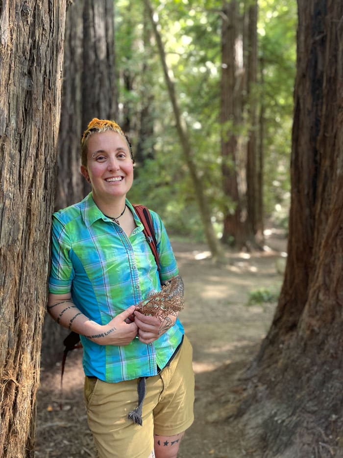 A person leans against a tree trunk, wearing a blue and green shirt.
