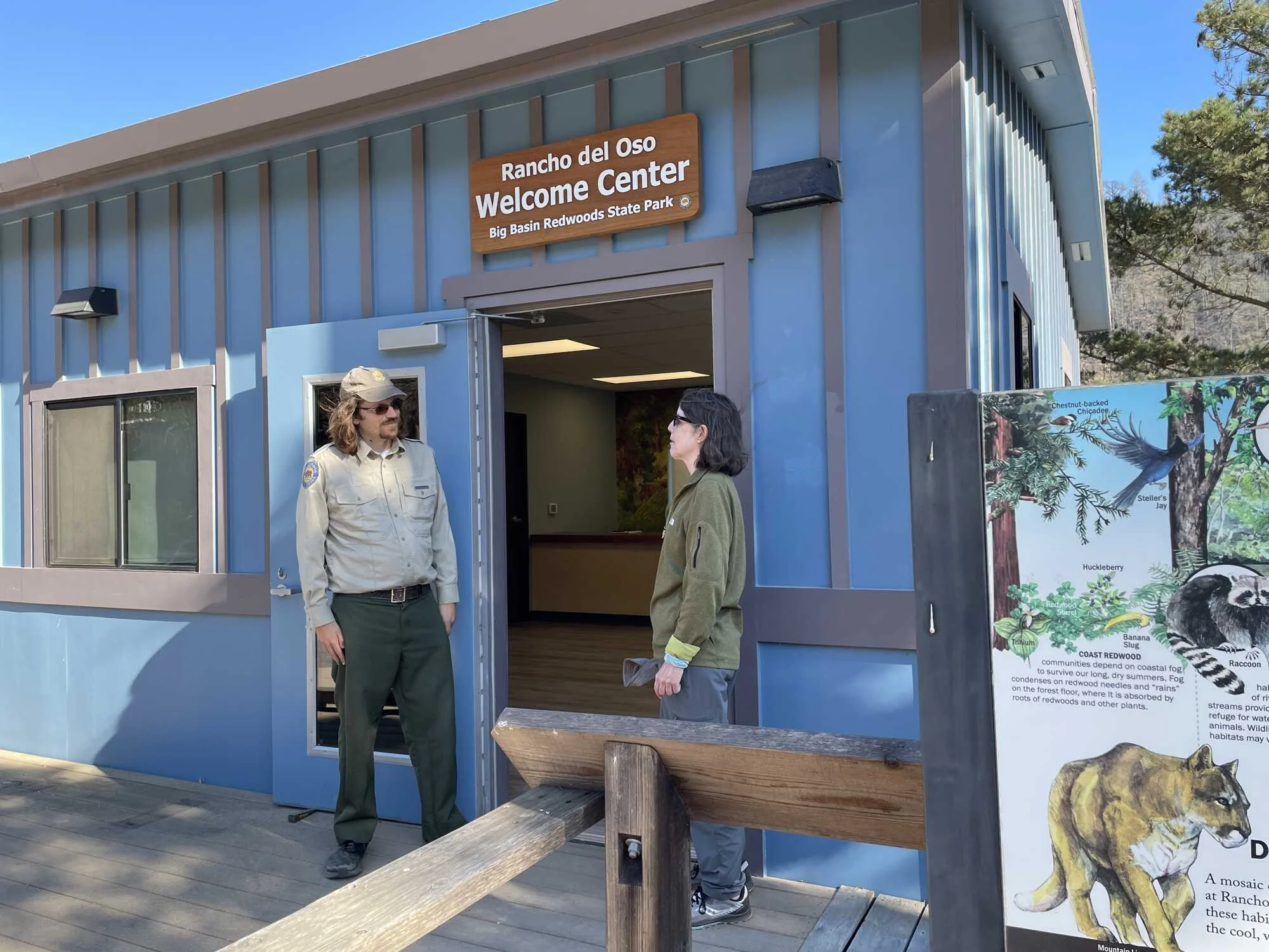 A ranger standing in front of the new Rancho del Oso Welcome Center - a blue and brown building with interpretive signage in front.