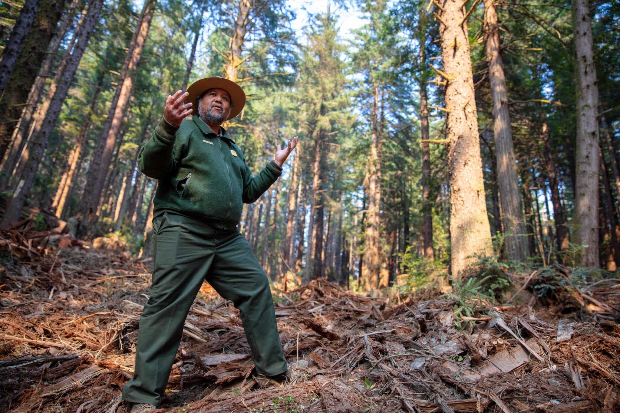 a man inb a green jumpsuit with a wide brimmed hat gestures while speaking in the middle of a forest clearing with lots of woody debris