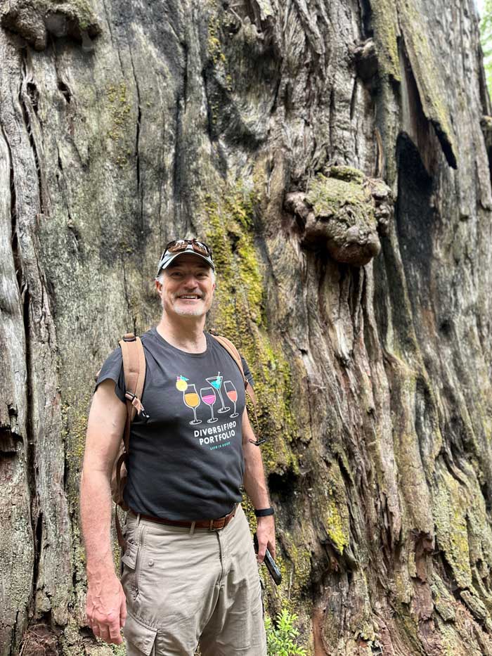 A man wearing a cap and a black t-shirt poses in front of a huge tree trunk