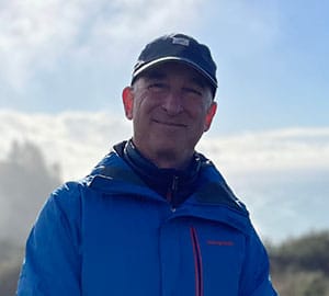 A white man wearing a hat, jeans and blue Patagonia jacket standing outside on a sunny day with redwoods and the ocean behind him.