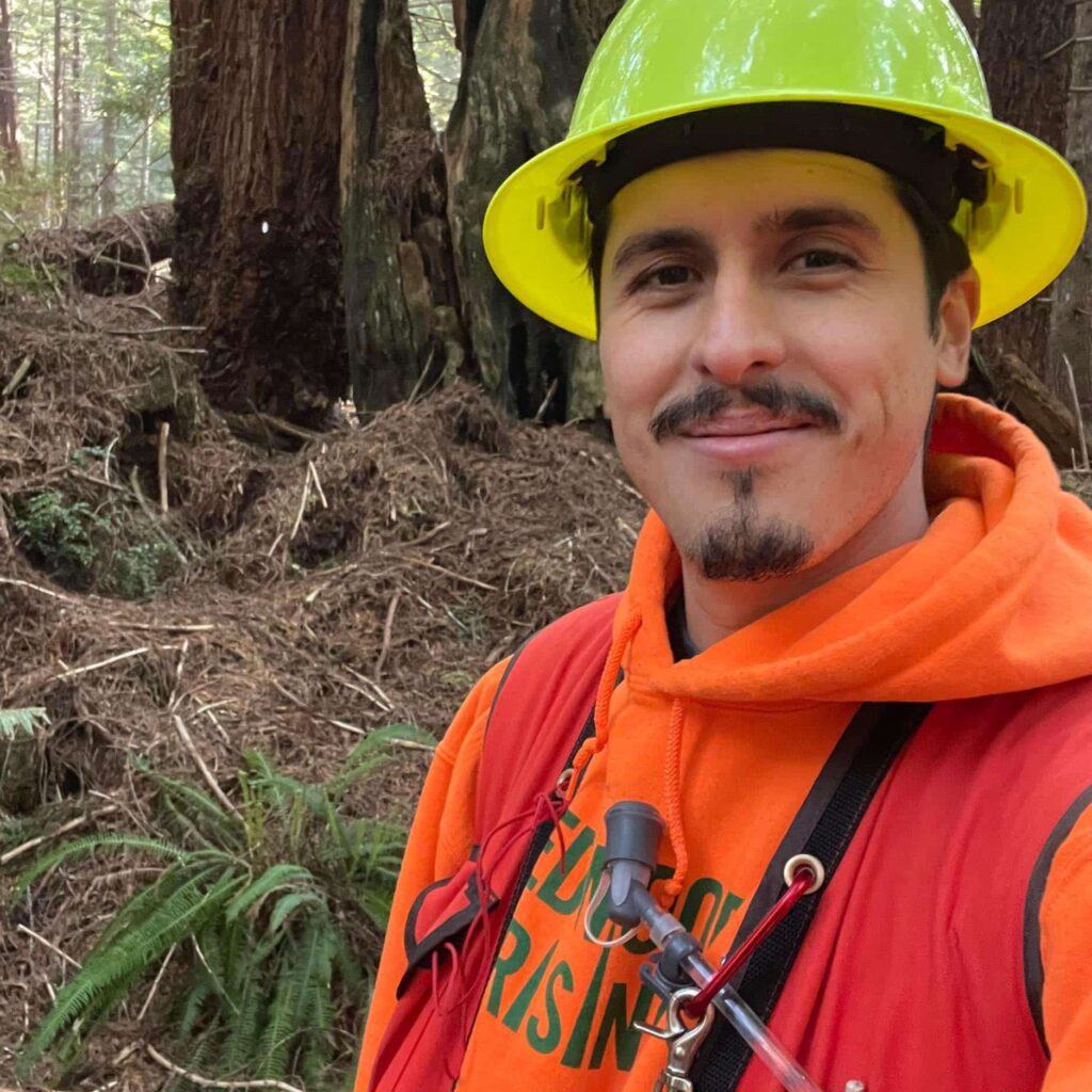 A close up selfie of a young man of Yurok heritage with a goatee wearing a yellow construction hat, orange Redwoods Rising hoodie, red vest with a camelback attachment standing in a redwoods forest.