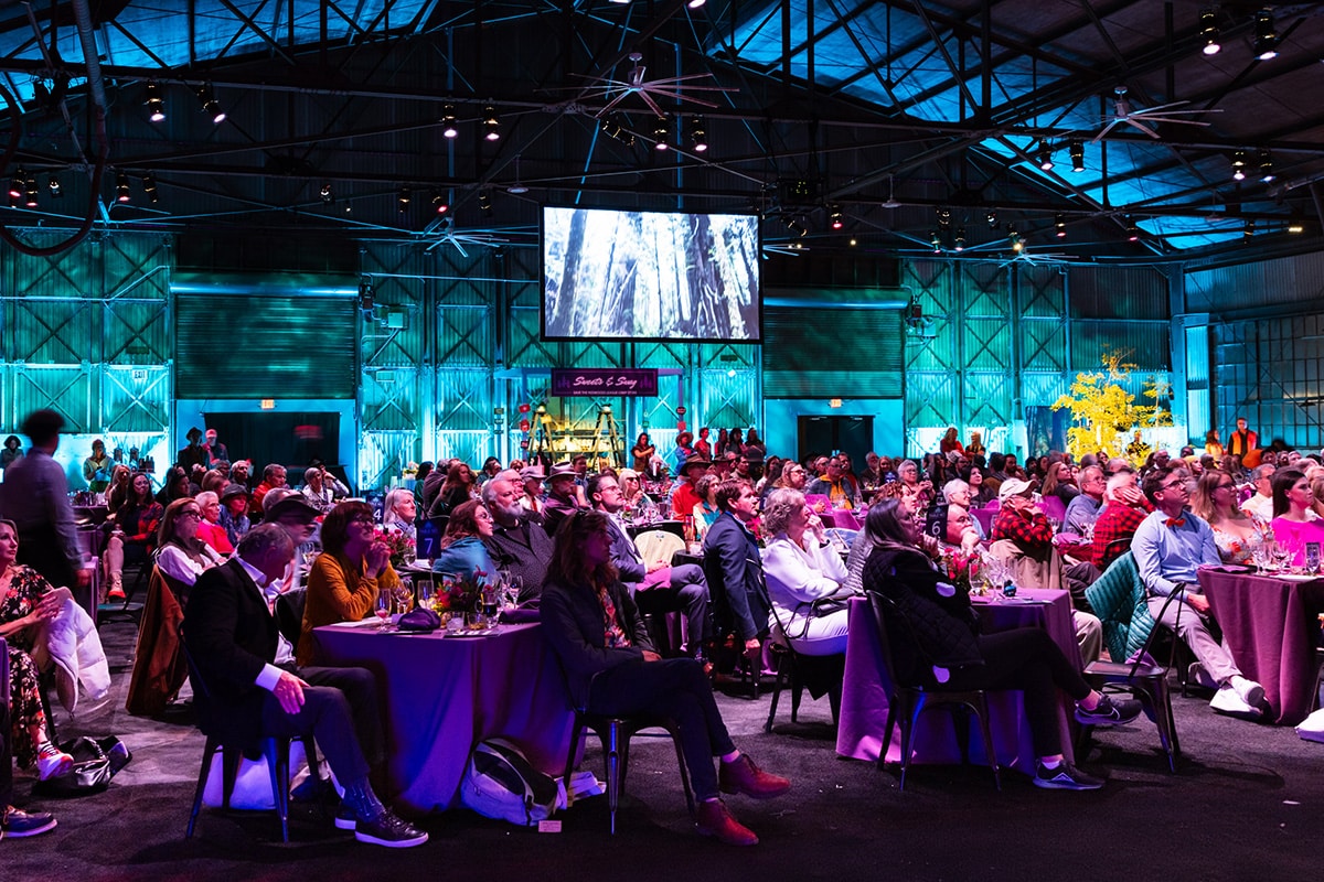 Many people seated around dining tables in the evening inside a decommissioned airplane hangar