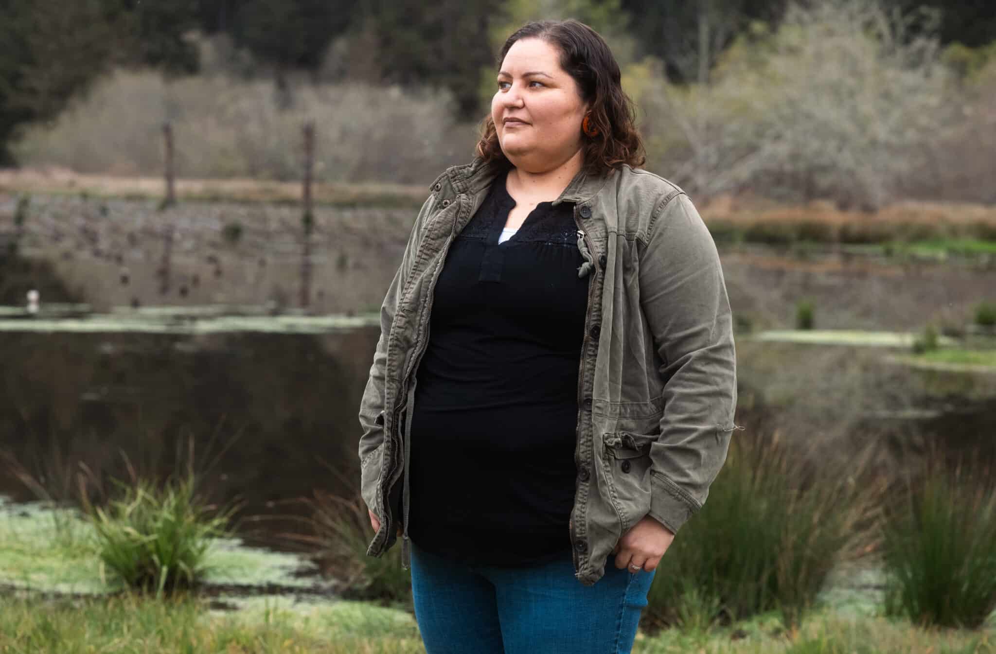a woman with wavy hair stares off to the left against a backdrop featuring a river and surrounding vegetation