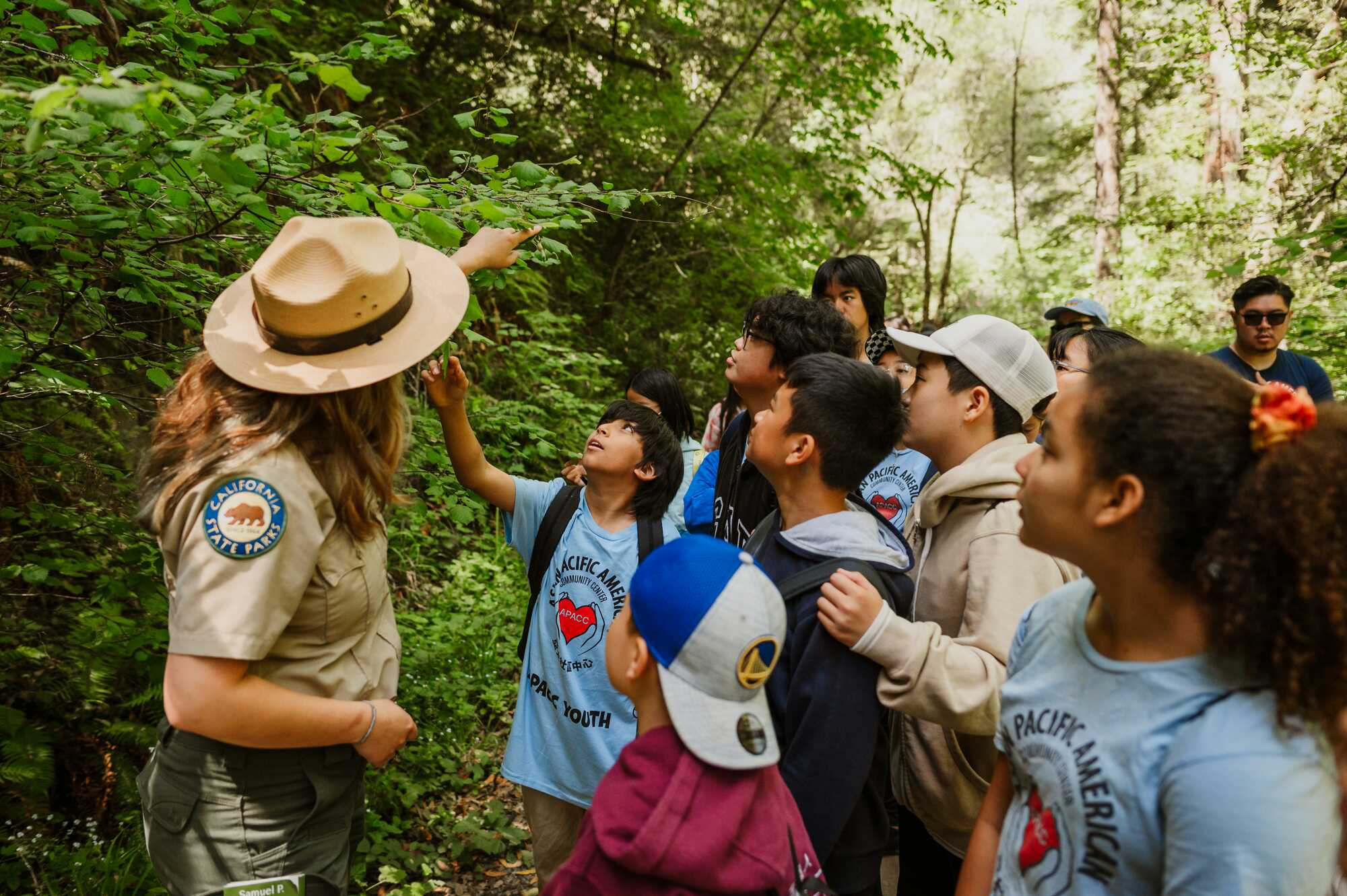 A group of school children look at and touch foliage along a trail with a state park ranger in the foreground.