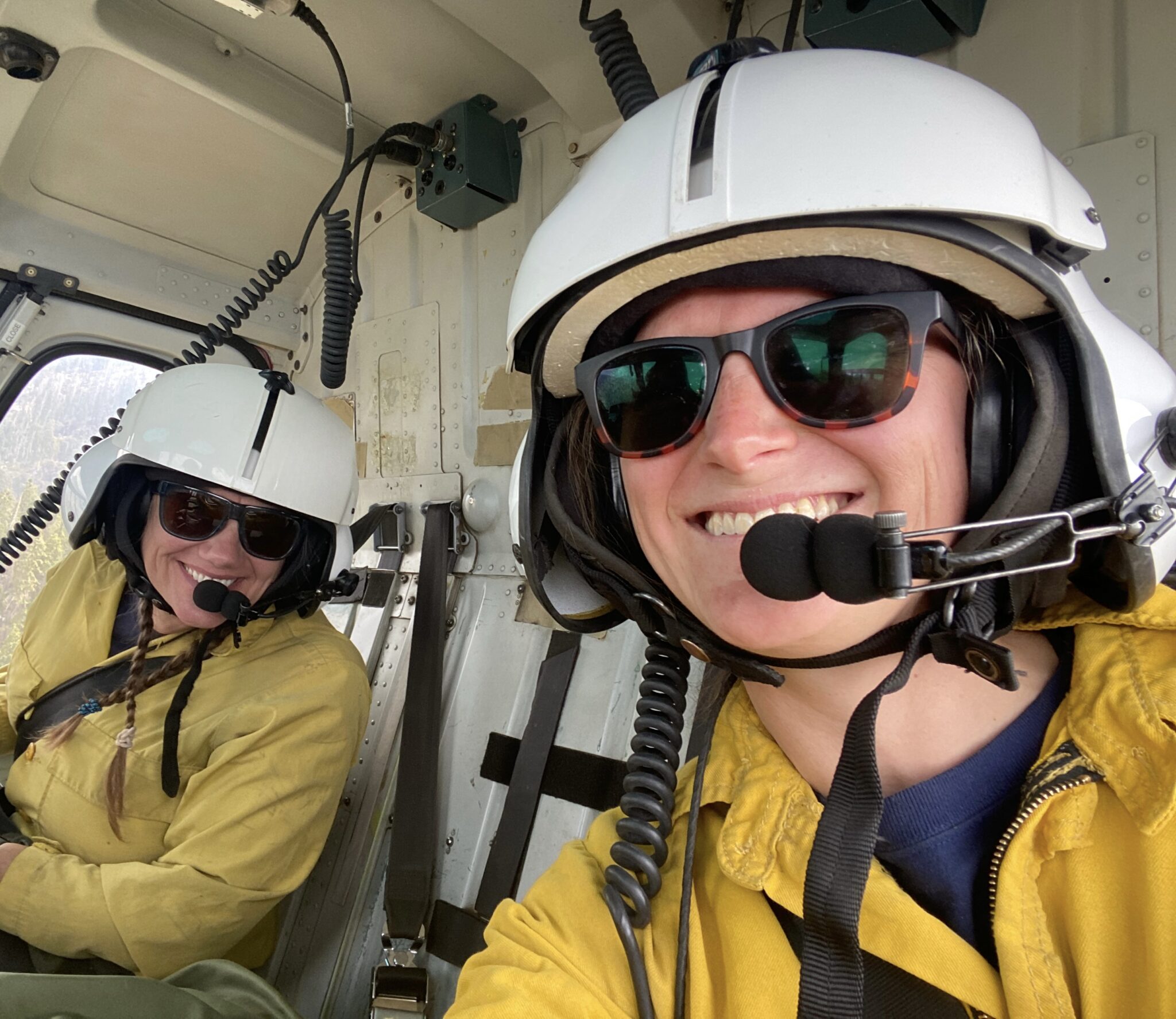 Two smiling women in a helicopter wearing white helmets and bright yellow safety jackets.