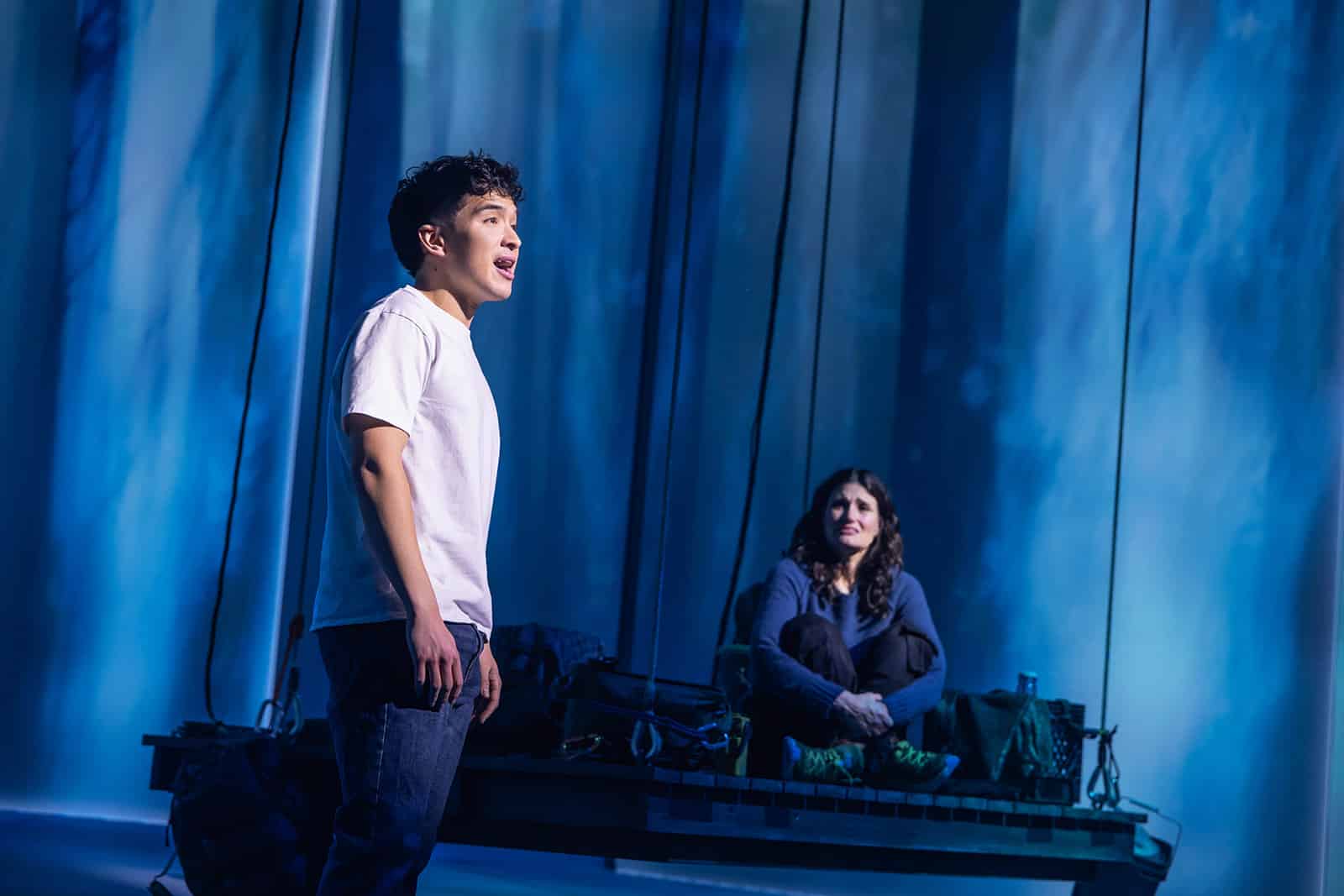A man stands the front of a stage in a white T-shirt, speaking or singing, while a woman sits on a platform in the background. The scene is illuminated with a cool blue forest backdrop.