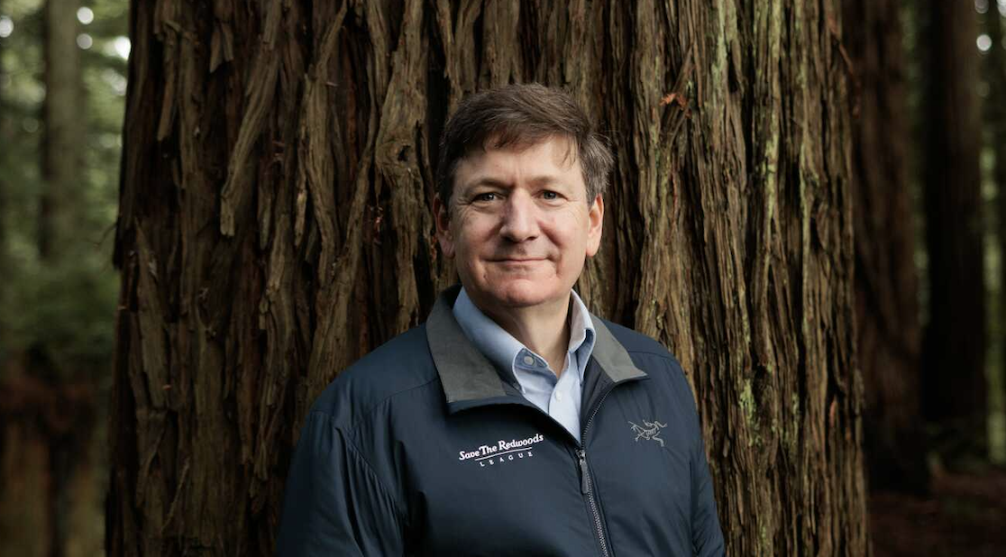 A man wearing a zipped jacket and blue shirt stands in front of a large tree trunk