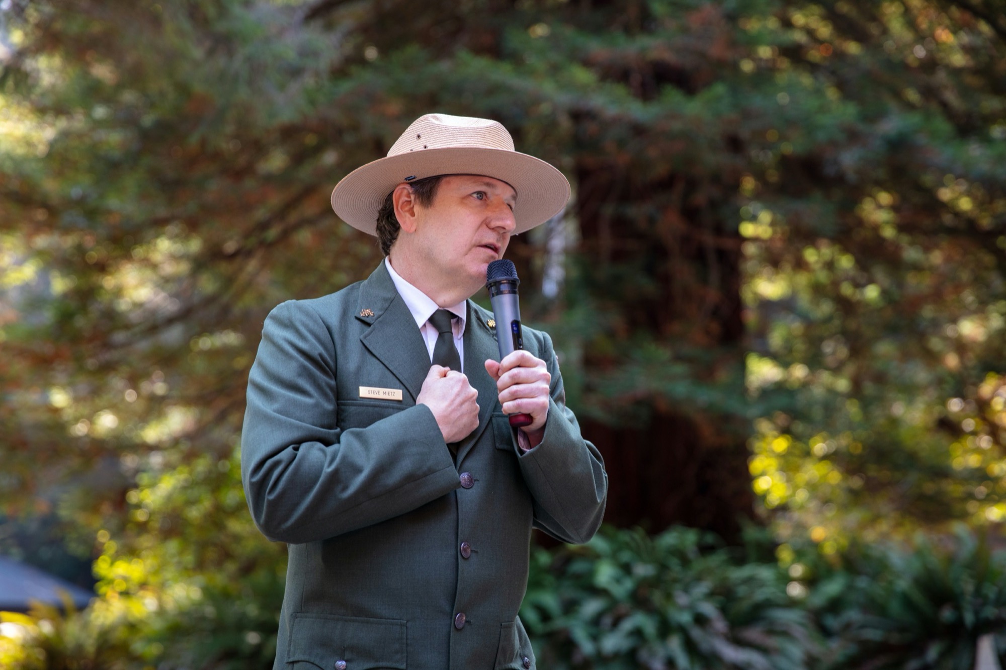 A man wearing a sun hat and a green blazer is speaking into a mic