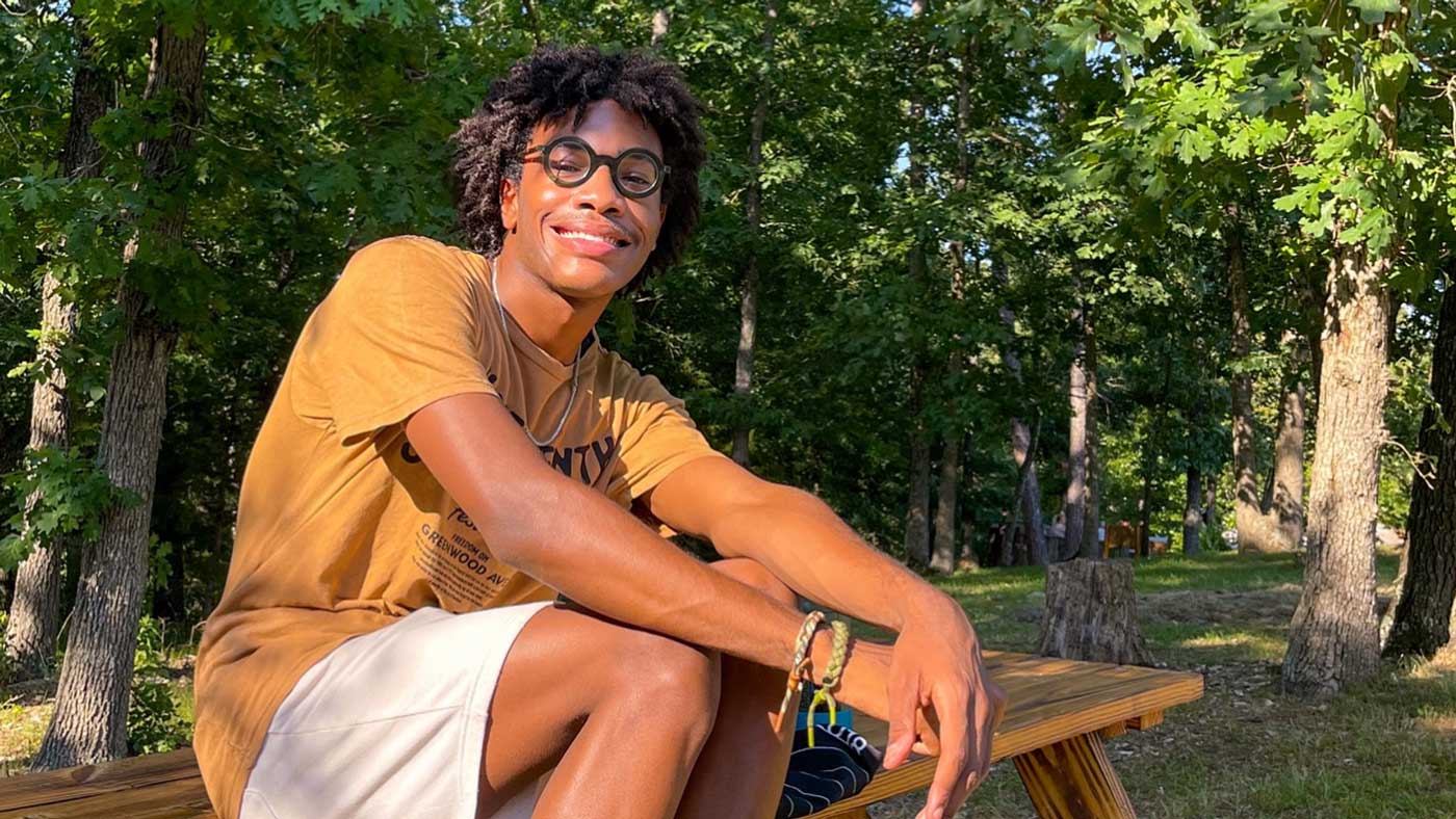 A young black man wearing round thick-framed glasses, a tan t-shirt, khaki shorts, and hemp bracelets smiles as he sits on a picnic table in a forested area outdoors.