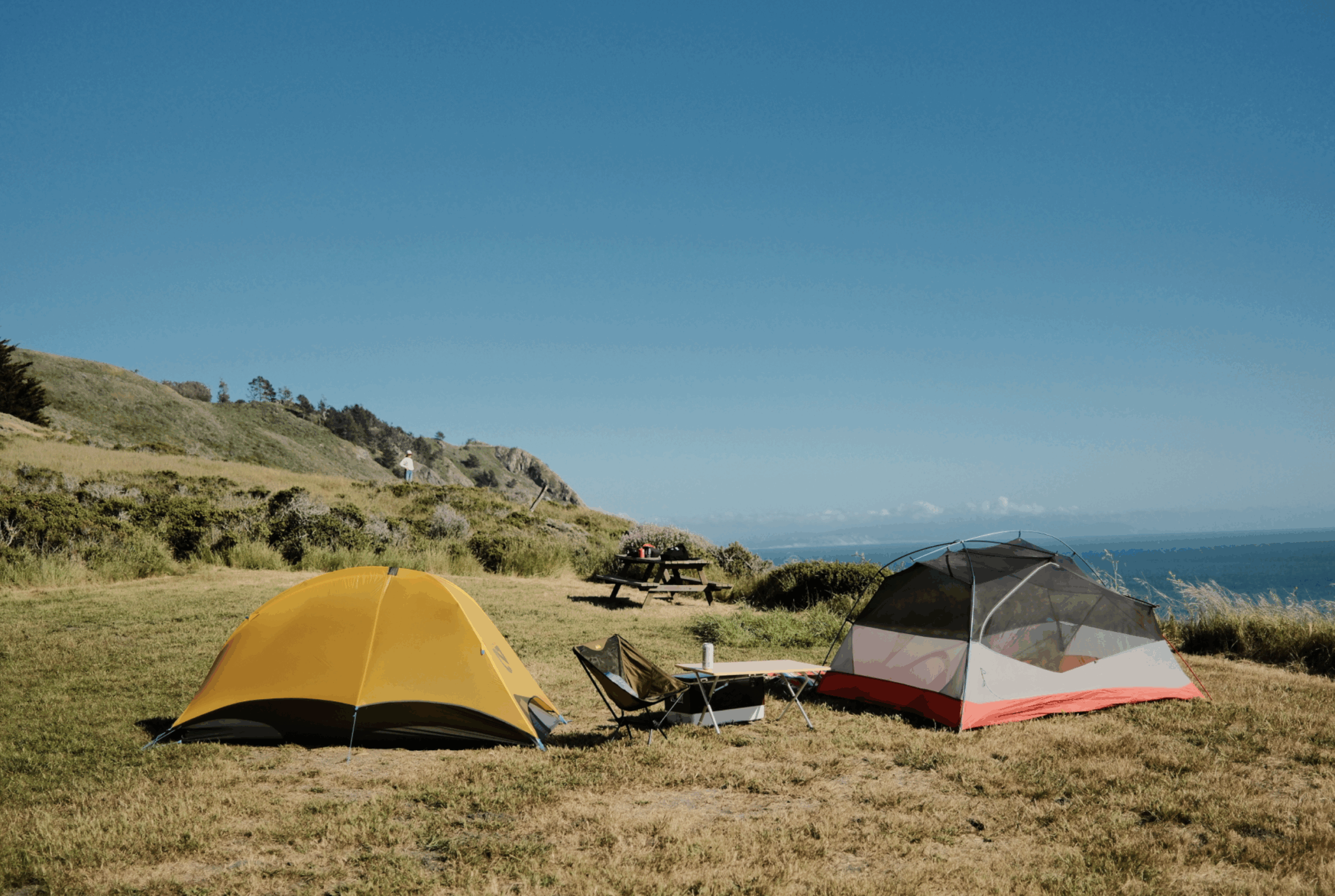 Two tents on a green plateau with a grassy hillside and blue skies in distance.