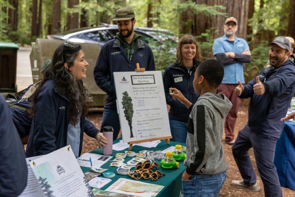 A group of people stand laughing around a table in a redwood forest.