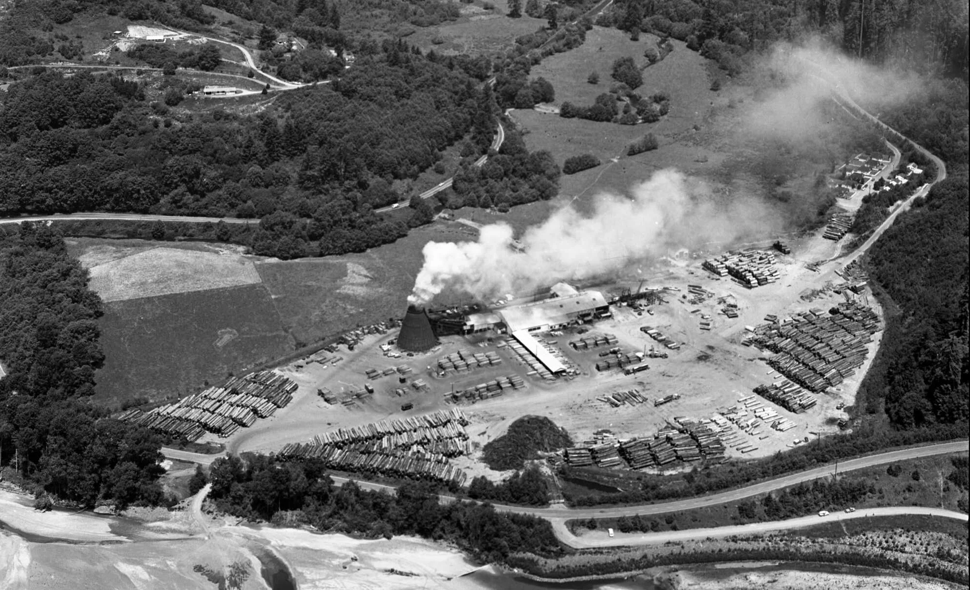 black and white image shows a sawmill with numerous logs and a smokestack