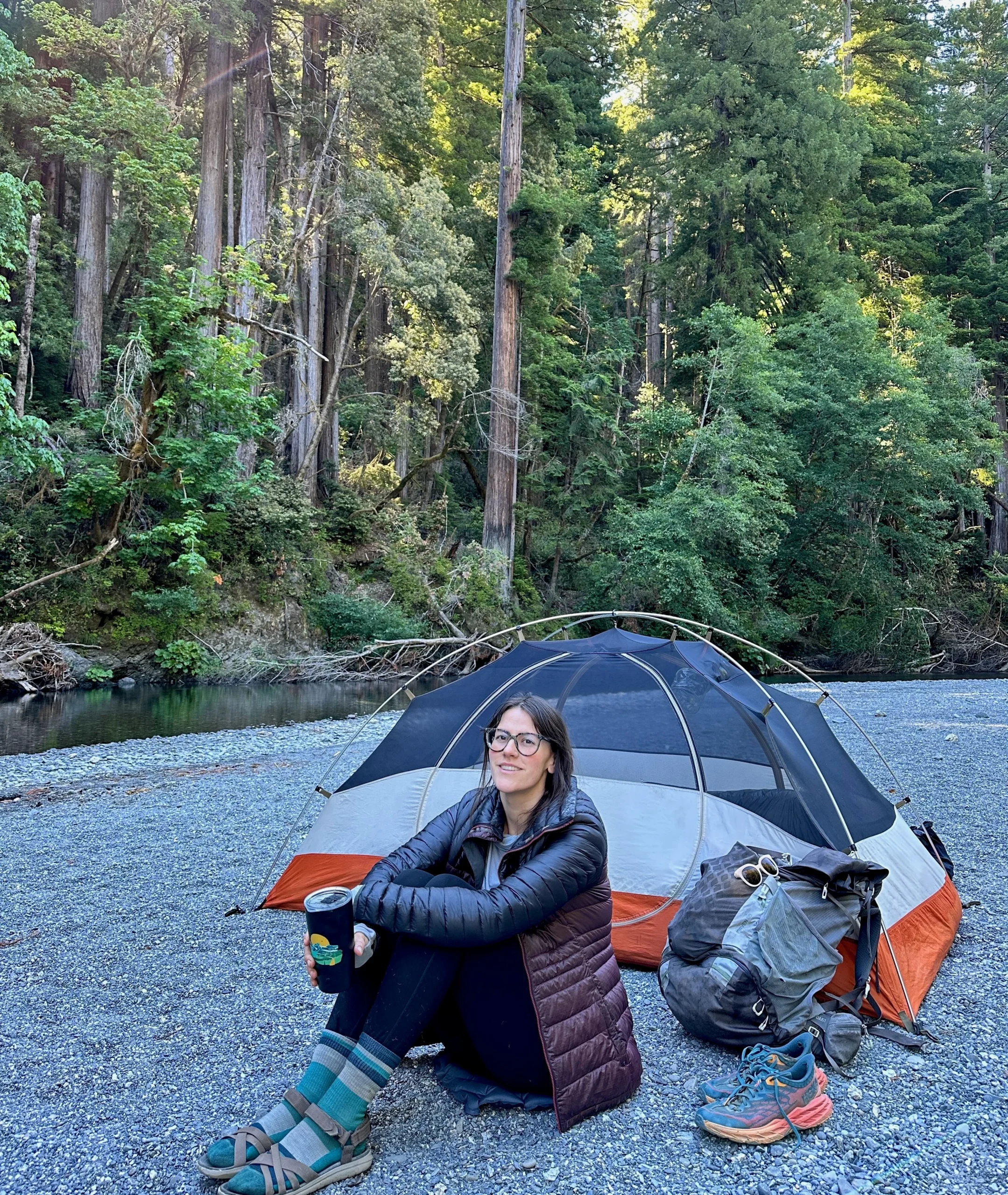 A woman sits in front of a tent on a creekside gravel bar.