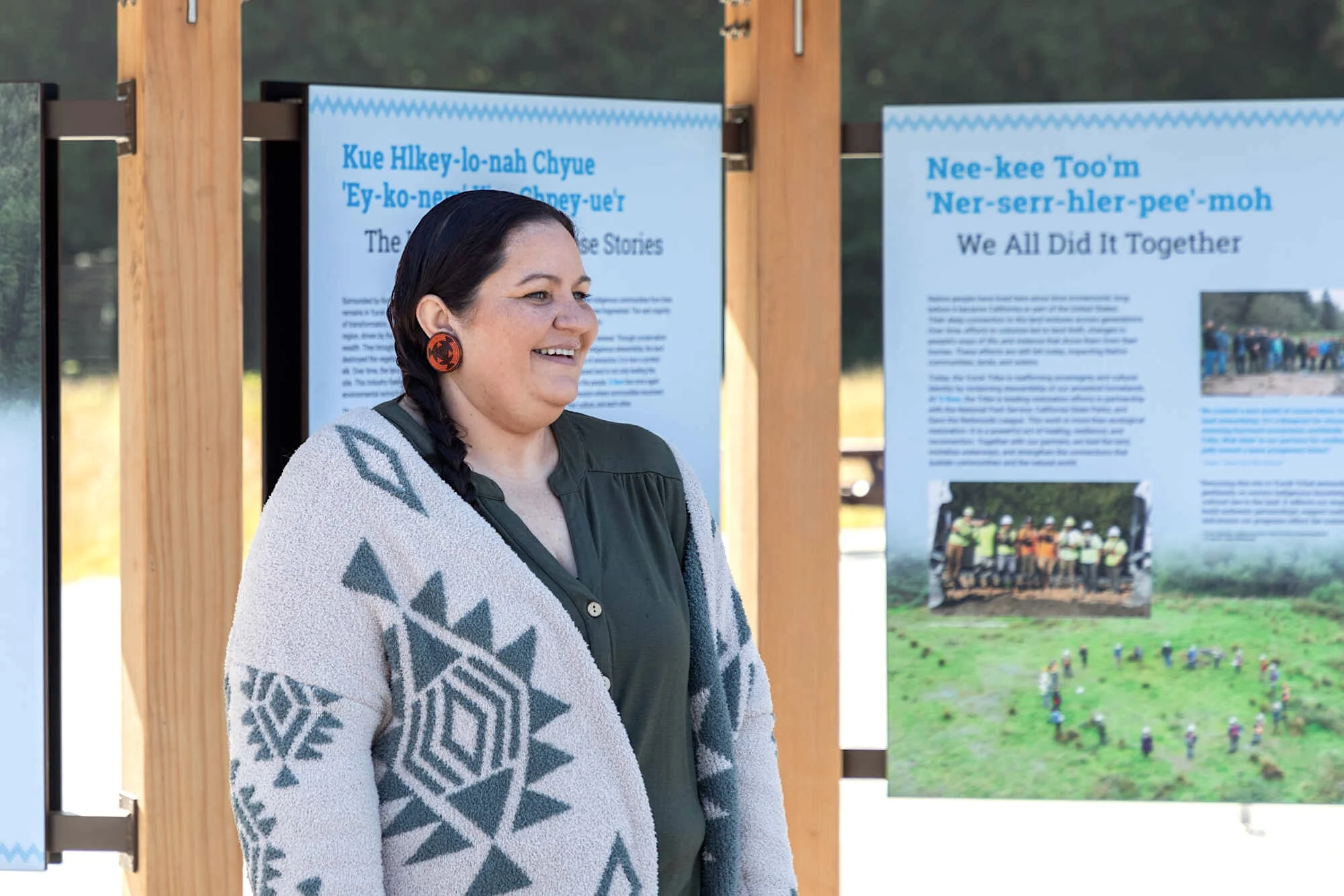 a smiling Yurok woman stands in front of welcome signs in Yurok and English