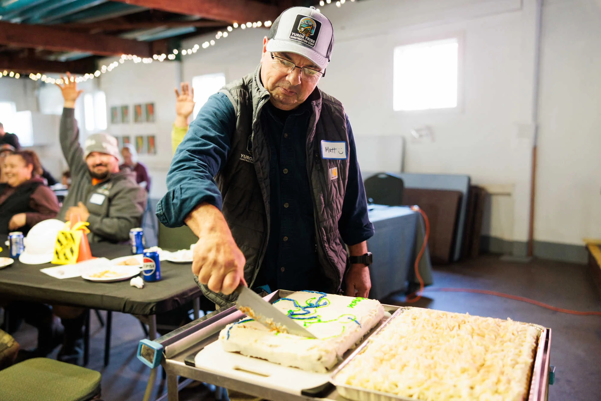 a man uses a large knife to slice a cake decorated with a blue stream and green riverbanks