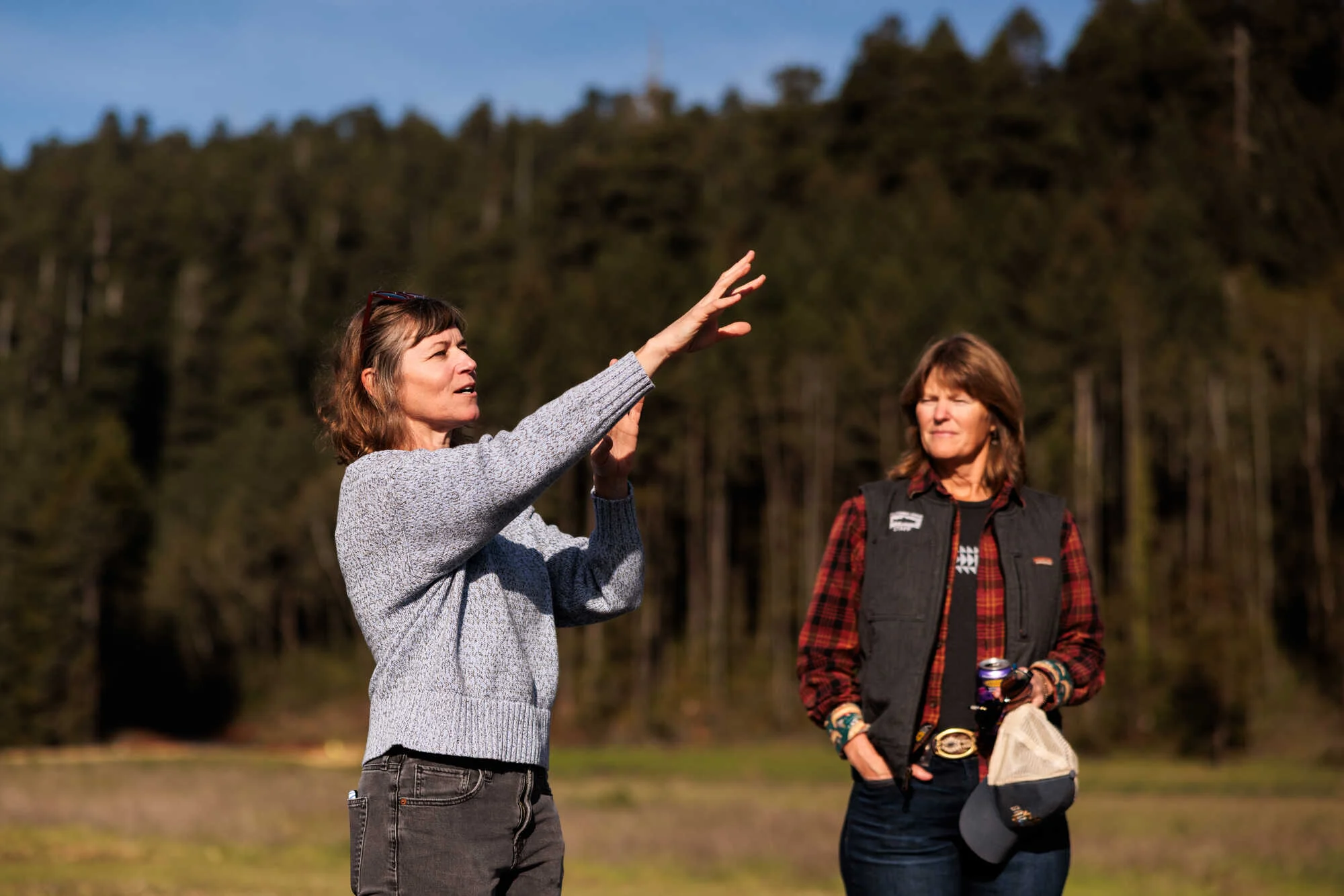 two woman stand in front of a redwood forest and one is using her raised hands to describe something.