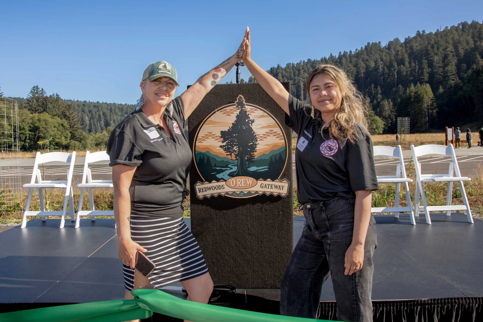 Two women high five over the podium with the logo for the new redwoods gateway, which depicts a massive tree against a tribal pattern on a river bank with a forest and mountain vista.