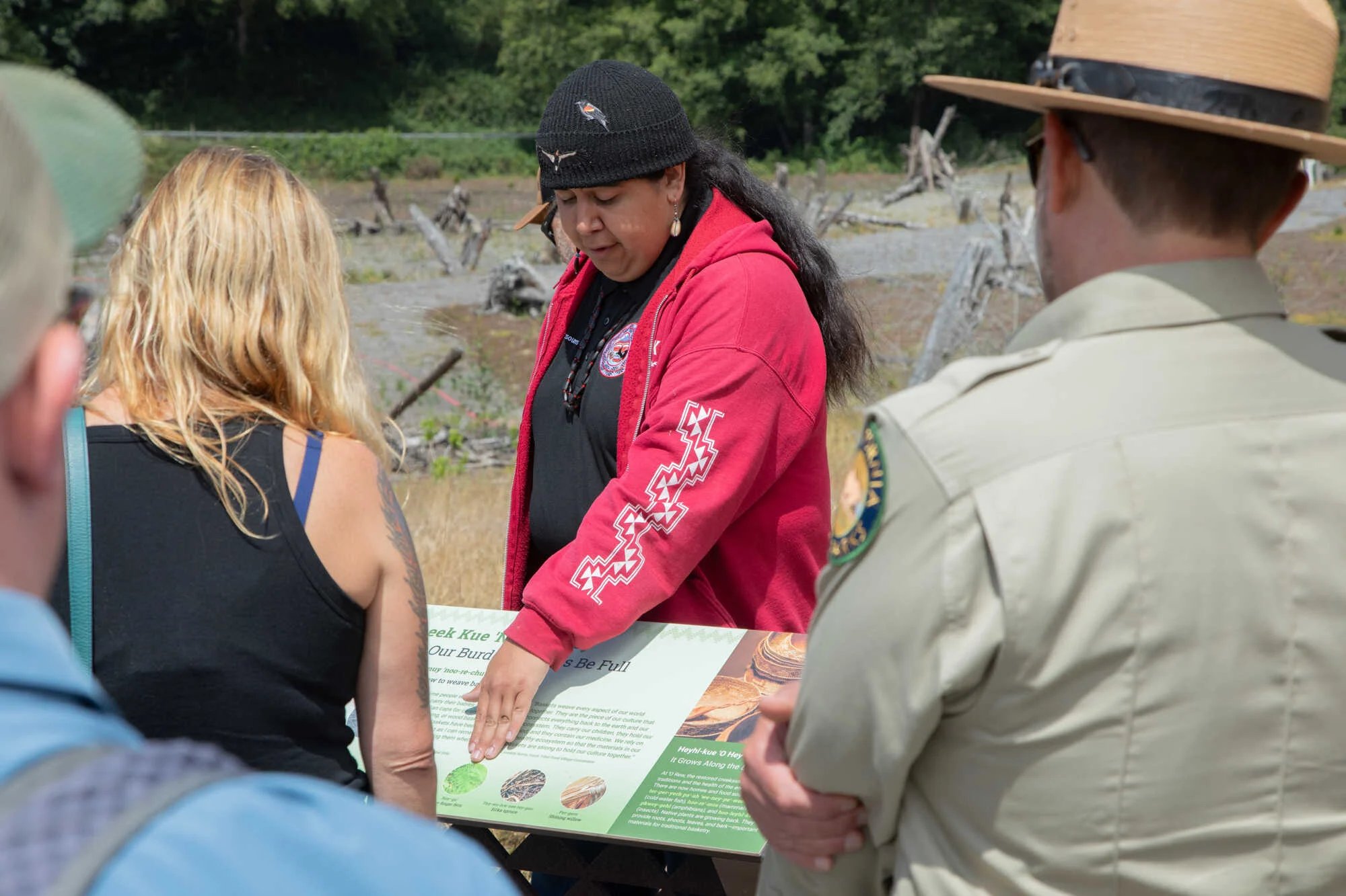 Indigenous man from the Yurok Tribe wearing a black beanie and red hoodie pointing at signage